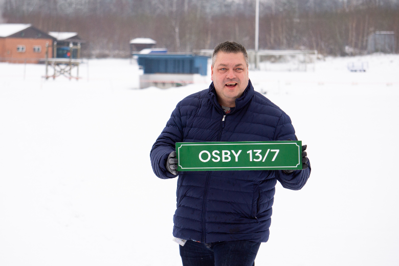 Jimmy står på ett snötäckt Osby idrottsplats med en skylt i handen som står Osby 13/9