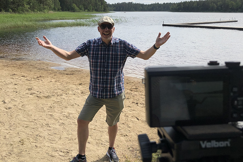 Kultur- och fritidschef Fredrik Johnsson står framför Hjärtasjön i Lönsboda en solig dag och sträcker ut armarna. Framför honom står en kamera på ett stativ. 