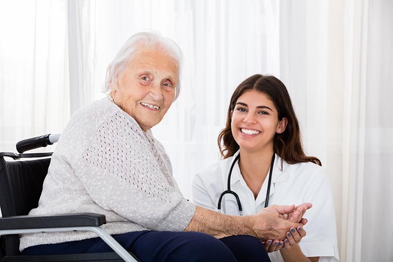 Glad äldre kvinna sitter i rullstol och ler mot kameran. Bredvid henne sitter en kvinnlig sjuksköterska som också ler mot kameran, runt sjuksköterskans hals sitter ett stetoskop. 