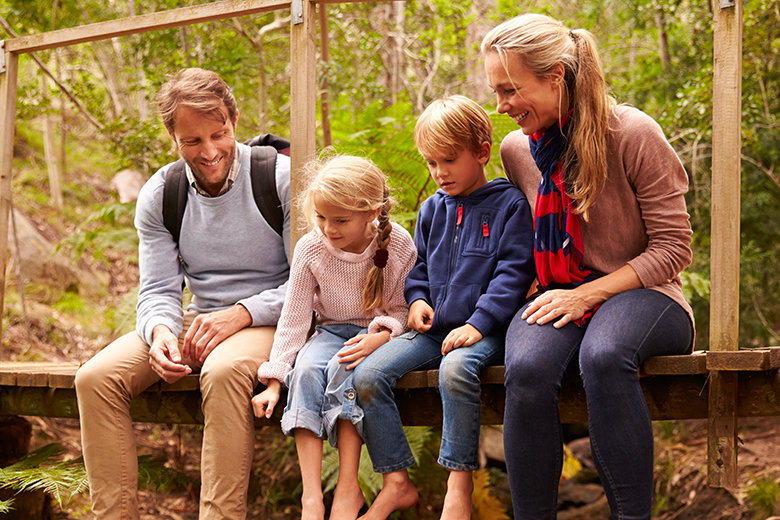 Glad familj med mamma, pappa och två barn sitter på en träbro i en skog